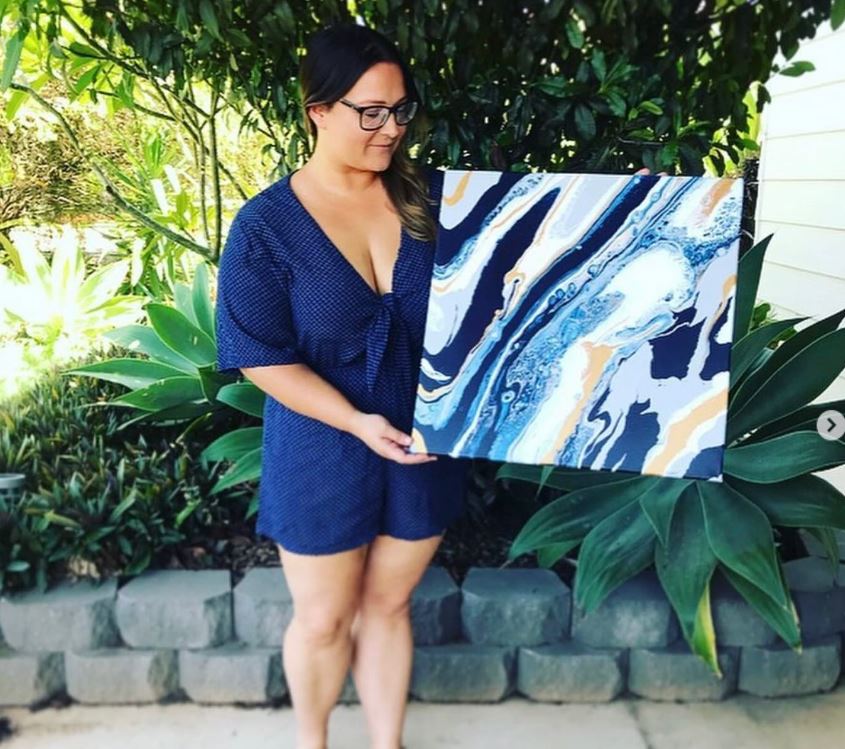 Woman in blue jumpsuit holds deep blue, white and gold fluid art on canvas piece in outdoor setting