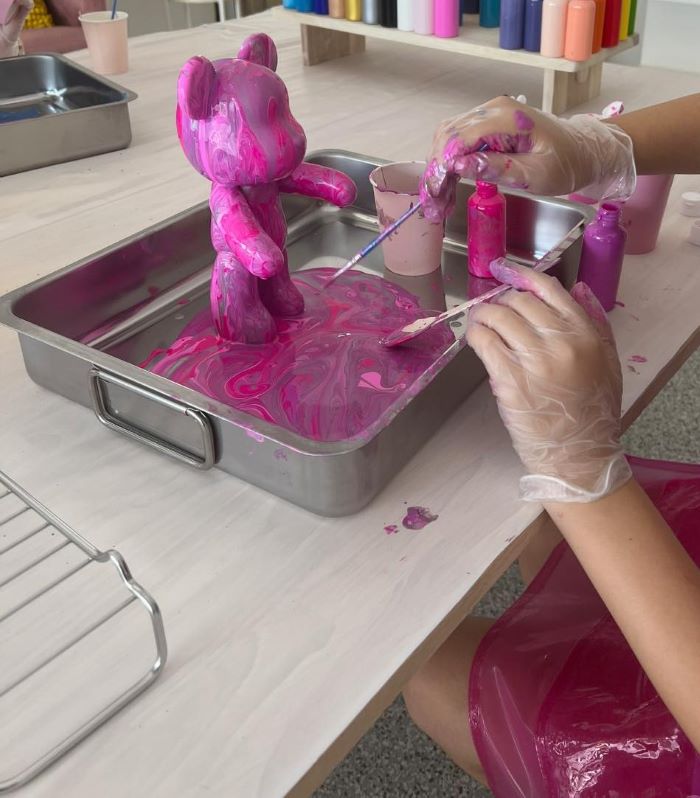 Fifth-Unicorn-Fluid-Bear-Art-Workshop-Customer-Girl2-cropped Fluid Bear Art Workshop using pinks, whites, and purples for flow paints on fluid bear. Two hands with gloves are painting gaps in fluid pour on bear. Fifth Unicorn is located in Newport, North Brisbane and runs creative art workshops, private events and posts Australia wide DIY art kits.