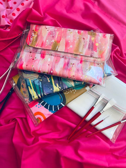 paint-a-clutch-3piece-workshop Painted handmade clutches with wrist straps in variety of colours and patterns, and blank unpainted clutch with paintbrushes on pink fabric background. Paint A Clutch Workshops by Fifth Unicorn are run at our Newport boutique art studio