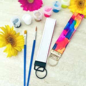 Handmade DIY Key Fob Wristlet - left is blank, right is painted with bright colours - Kit by Fifth Unicorn with 2 paintbrushes, 6 paint colours in mini pot set of yellow, blue, pink, gold, white and teal, with 3 flowers as decorative items. Fifth Unicorn holds creative Art workshops in Newport, near Redcliffe, Brisbane