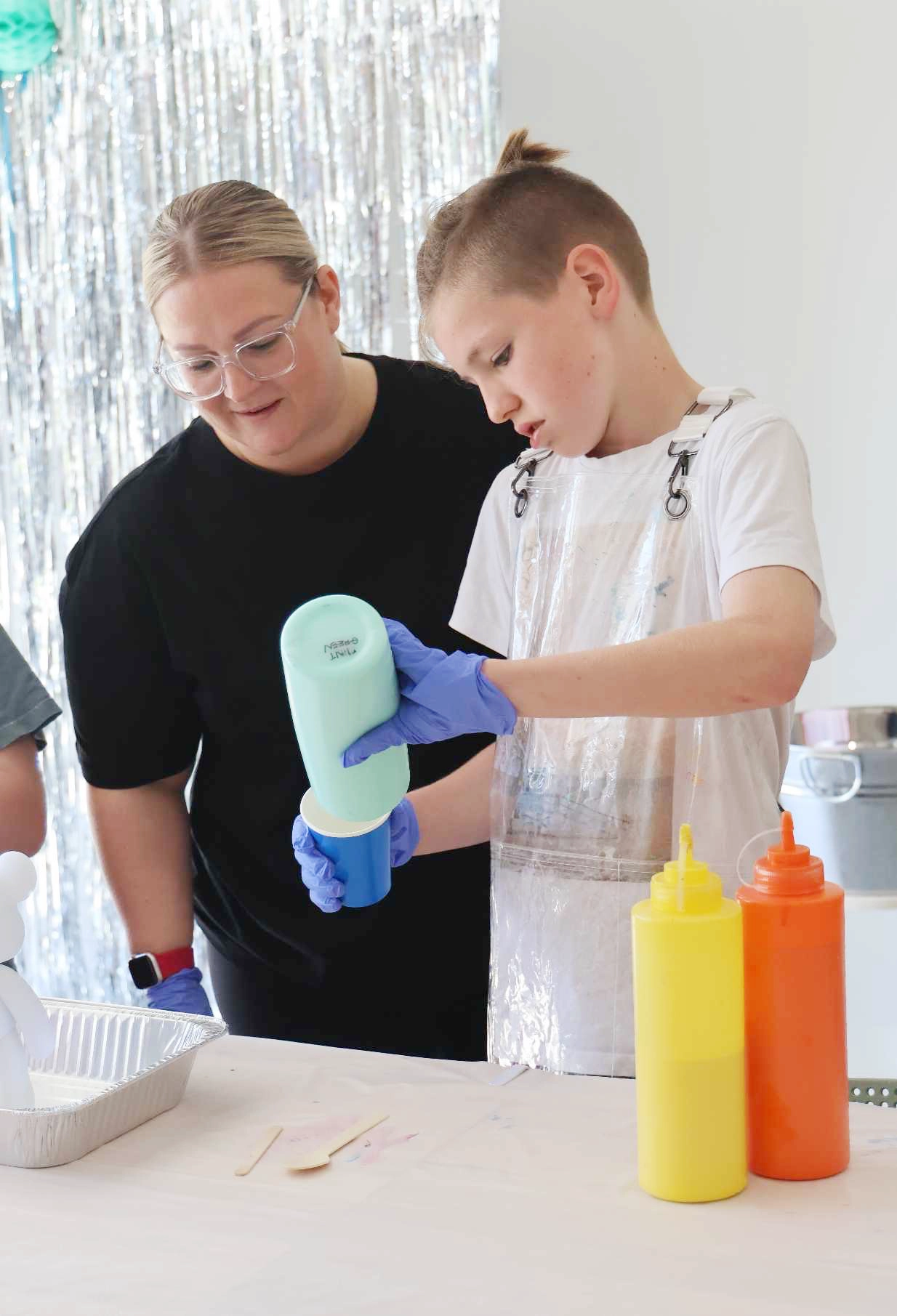 A child wearing a clear apron and blue gloves is pouring light blue paint into a cup, while an adult watches and supervises closely. Three bottles of brightly coloured paint—yellow, orange, and red—are on the table next to them, along with a metal tray and wooden sticks. Both are focused on the creative activity in a light-filled room decorated with shiny silver streamers.