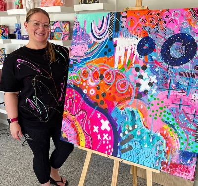 A woman wearing glasses and a black shirt with colourful splatters stands smiling next to a large, vibrant abstract painting on an easel. The painting features bold colours such as pink, blue, orange, and white with various shapes, squiggles, dots, and crosses. Shelves with other colourful artwork are visible in the background.