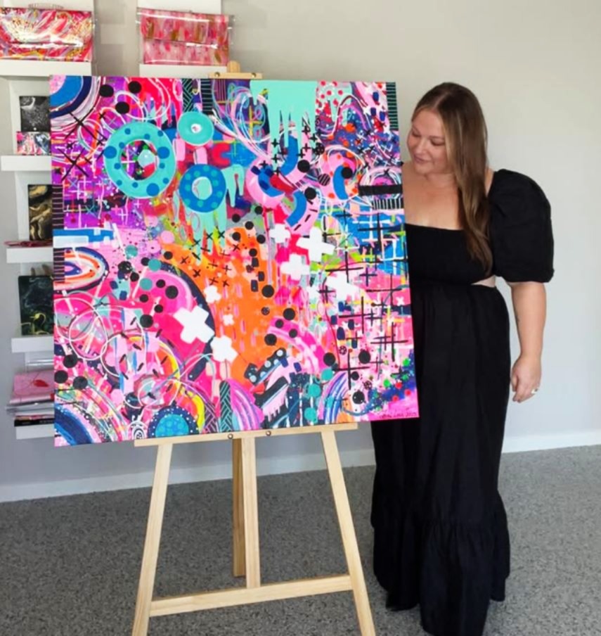A woman in a black dress stands beside a large, colourful abstract painting on an easel. The artwork is vibrant, featuring a mix of neon pinks, oranges, blues, and greens, with bold shapes, dots, lines, and crosses. The woman is smiling, looking proudly at the painting. The room is well-lit with neutral walls, and there are shelves with more art and supplies in the background.
