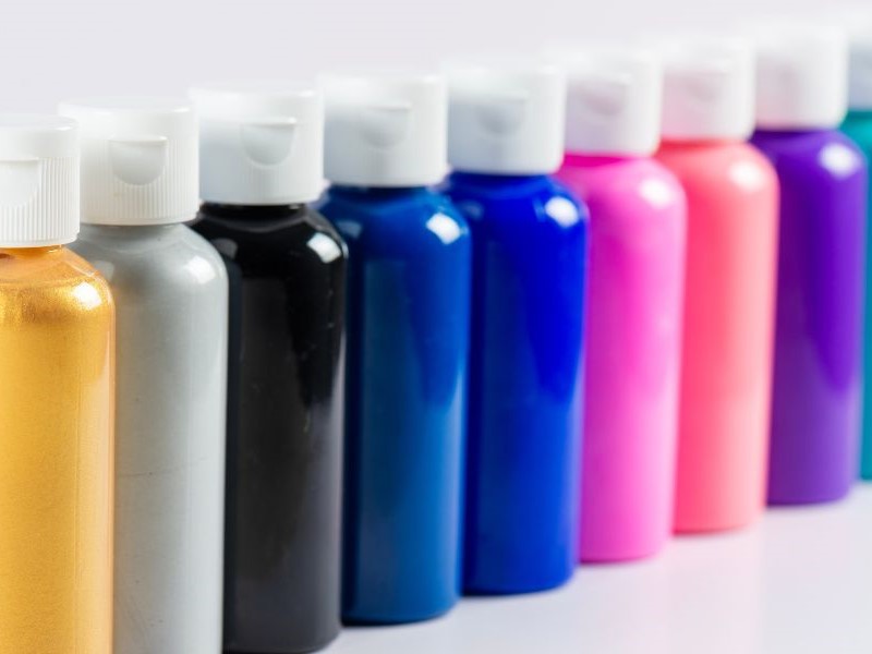 A row of colourful plastic bottles with white flip-top caps, containing different shades of paint or craft materials, arranged side by side from gold, silver, black, blue, purple, pink, red, and other colours.