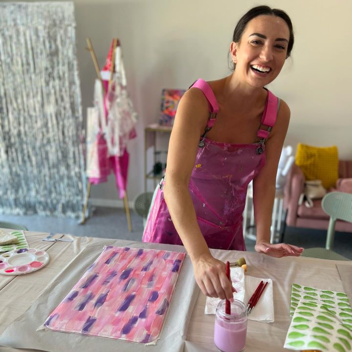 A woman wearing a pink paint-splattered apron is smiling and standing at a table set up for painting. She is dipping a paintbrush into a jar of light purple paint. On the table are a palette, painted sheets of paper, and art supplies. The background shows a studio space with hanging aprons, a lounge chair with a yellow cushion, and a shimmering curtain. The scene is bright and cheerful.